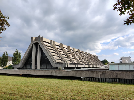 Außenansicht der Glaskathedrale. Dreieckige Giebelform aus hellgrauem Beton. Unten Rasen, oben Wolken