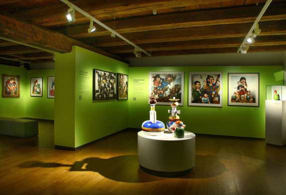 Blick in die Ausstellung von Michael Mathias Prechtl mit hellgrünen Wänden, daran Gemälde und drei bunte Tonfiguren in der Raummitte.