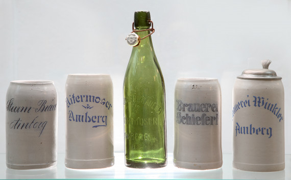 Vier graue Bierkrüge von verschiedenen Brauereien und eine grüne Bierflasche dazwischen.