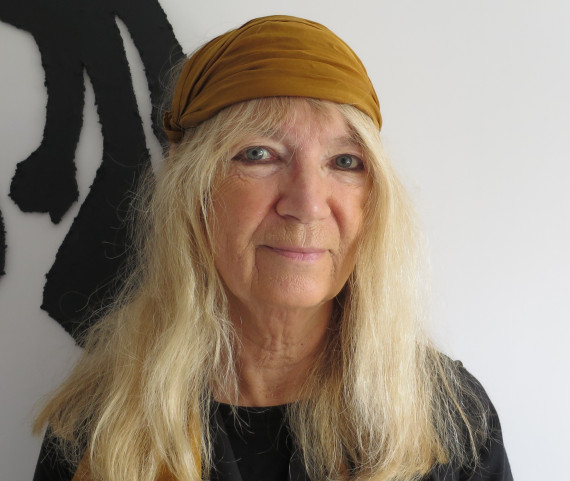 Frontales Brustbild der Künstlerin Petra Naumann vor einer weißen Wand mit schwarzen Mustern. Sie hat lange blonde Haare, trägt ein ocker-farbenes Haarband und ein schwarzes T-Shirt  und schaut freundlich.