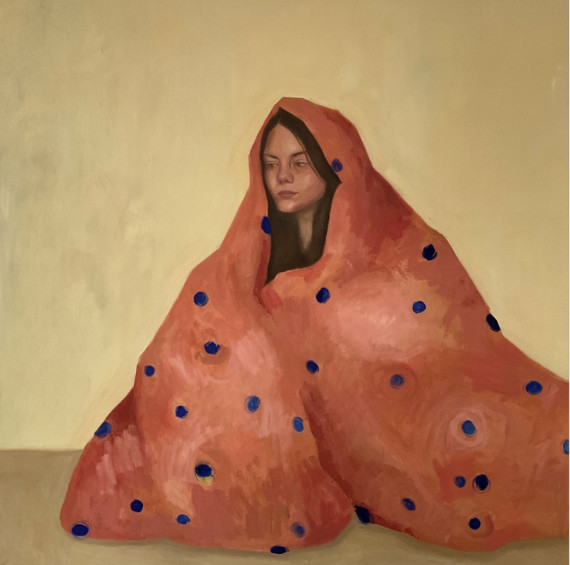 Selbstporträt der Künstlerin Jule Rdurof in sitzender Pose unter einer orangen Decke mit blauen Punkten in einem beigen, leeren Umraum.