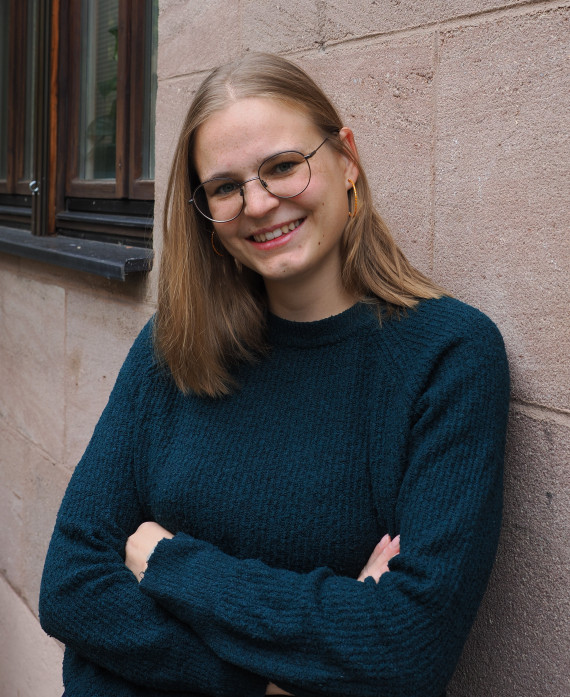 Hüftporträt der nach rechts an eine Wand gelehnten Künstlerin mit verschränkten Armen. Sie hat schulterlanges dunkelblondes Haar, trägt eine runde Brille mit dünner FAssung und einem dunkelblauen Pubblover. Sie lächelt.