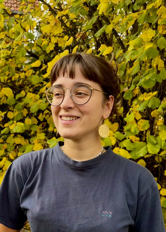 Porträtfoto von Kim Nespor. Brustbild mit der nach links blickenden Künstlerin. Sie hat dunkle, zurückgebundene Haare mit einem Pony, trägt eine runde Brille mit dünnem Gestell und ein dunkelblaues Shirt.