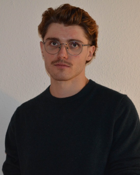 Porträtfoto vom Künstler vor einer weißen Wand. Brustbild des Künstlers mit kurzen Haaren, einer runden Brille mit dünnem Gestellt und einem schwarzen Pullover. Er blickt frontal in die Kamera.