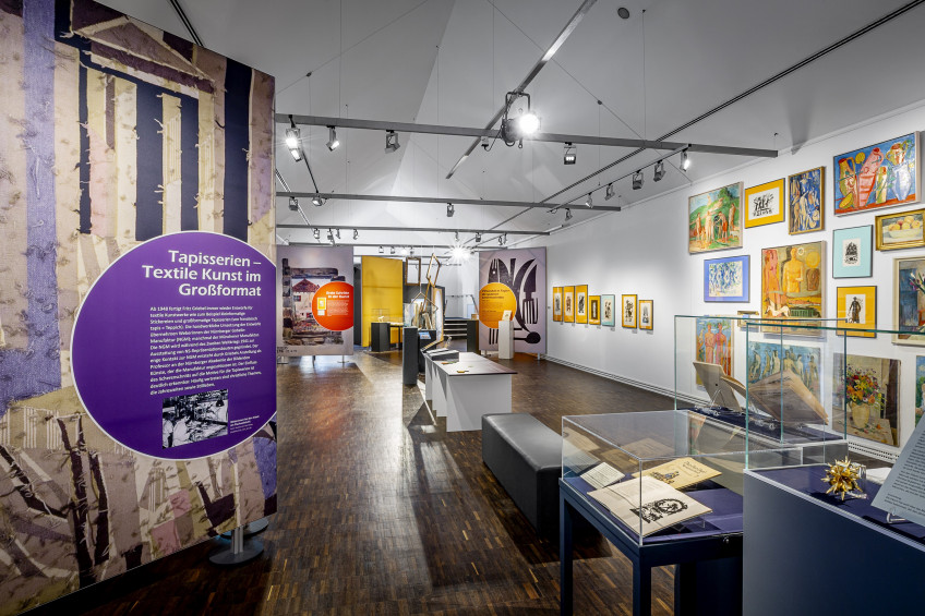 Blick in den länglichen Sonderausstellungsraum mit großen Textspanntafeln links sowie in der Raummitte Tischvitrinen, einer Sitzbank und einem Tisch.