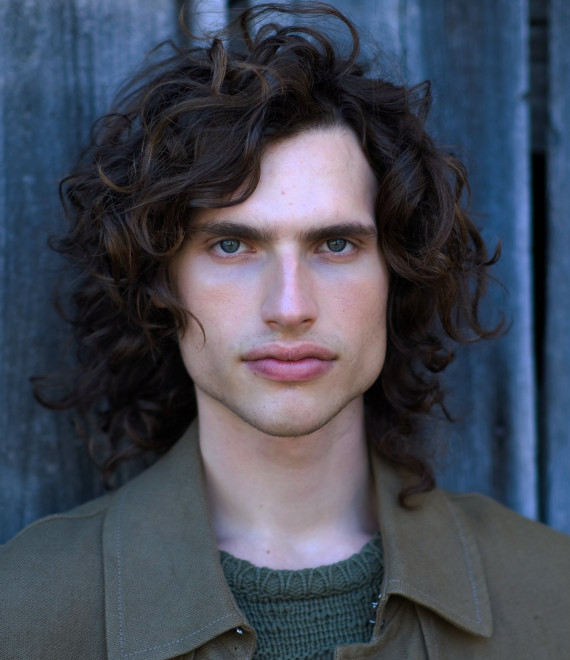 Frontales Portr&auml;t eines jungen Mannes mit lockigem, schulterlangen Haar und strengem Blick.