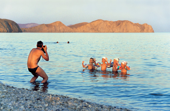 Foto eines im Wasser stehenden Mannes in Badehose, der vier kleine, winkende Kinder im Wasser vor sich fotografiert.