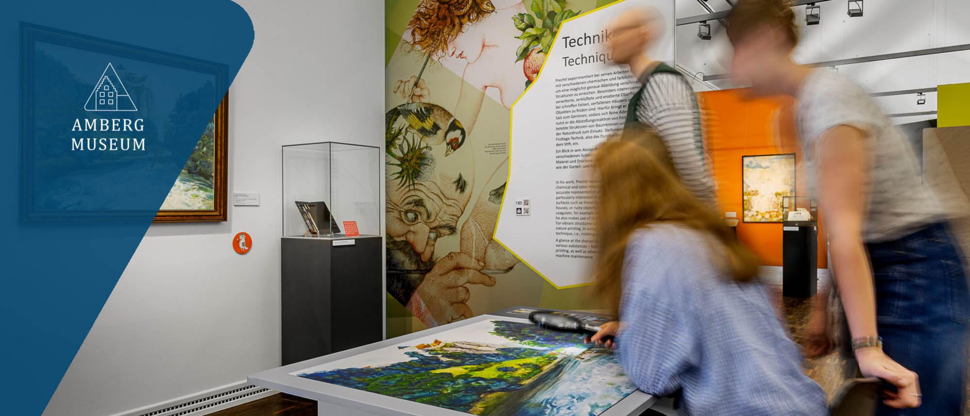 Zwei Menschen an einem Tisch mit einem bunten Bild . Sie schauen es durch eine Lupe genau an. Dahinter steht eine dritte Person, die Text an einer gro&szlig;en Text-Bild-Tafel liest. Daneben steht eine Vitrine. 