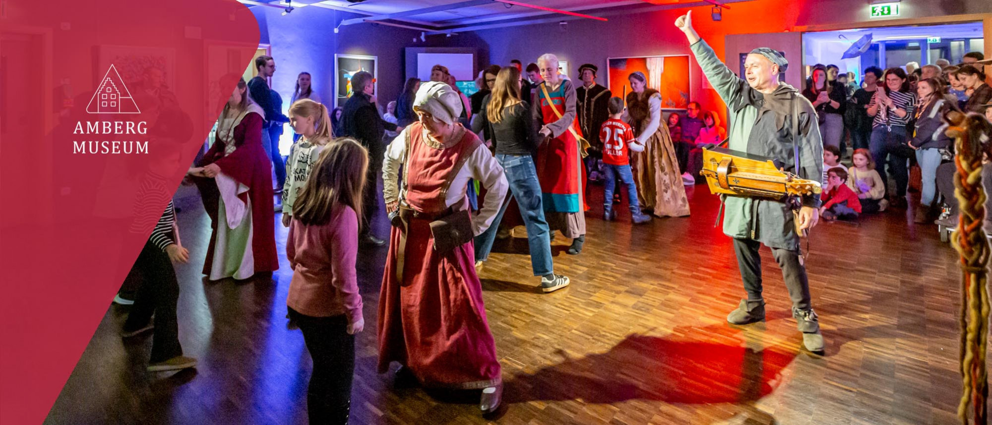 Menschen in historischer und moderner Kleidung Tanzen zusammen in einem bunt beleuchteten Raum im Museum. In der Mitte steht ein Mann mit einer historischen Leier.