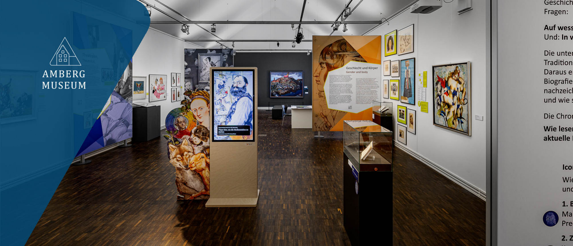 Blick in einen schmalen, tiefen Ausstellungsraum mit bunten Texttafeln, Vitrinen und einer hochformatigen Medienstehle in der Raummitte.